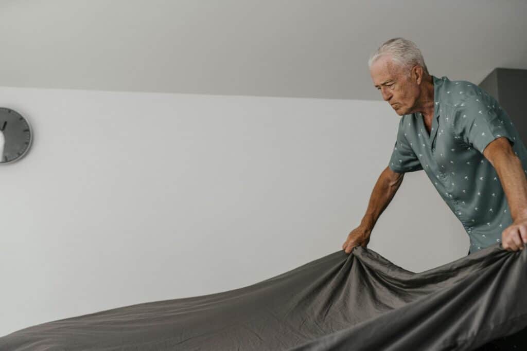 Elderly man with gray hair making bed in modern minimalist room.